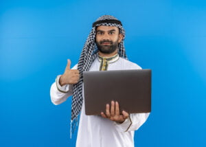 Menulis-Arab-Di-Word-arabic-businessman-traditional-wear-holding-laptop-smiling-showing-thumbs-up-standing-blue-wall